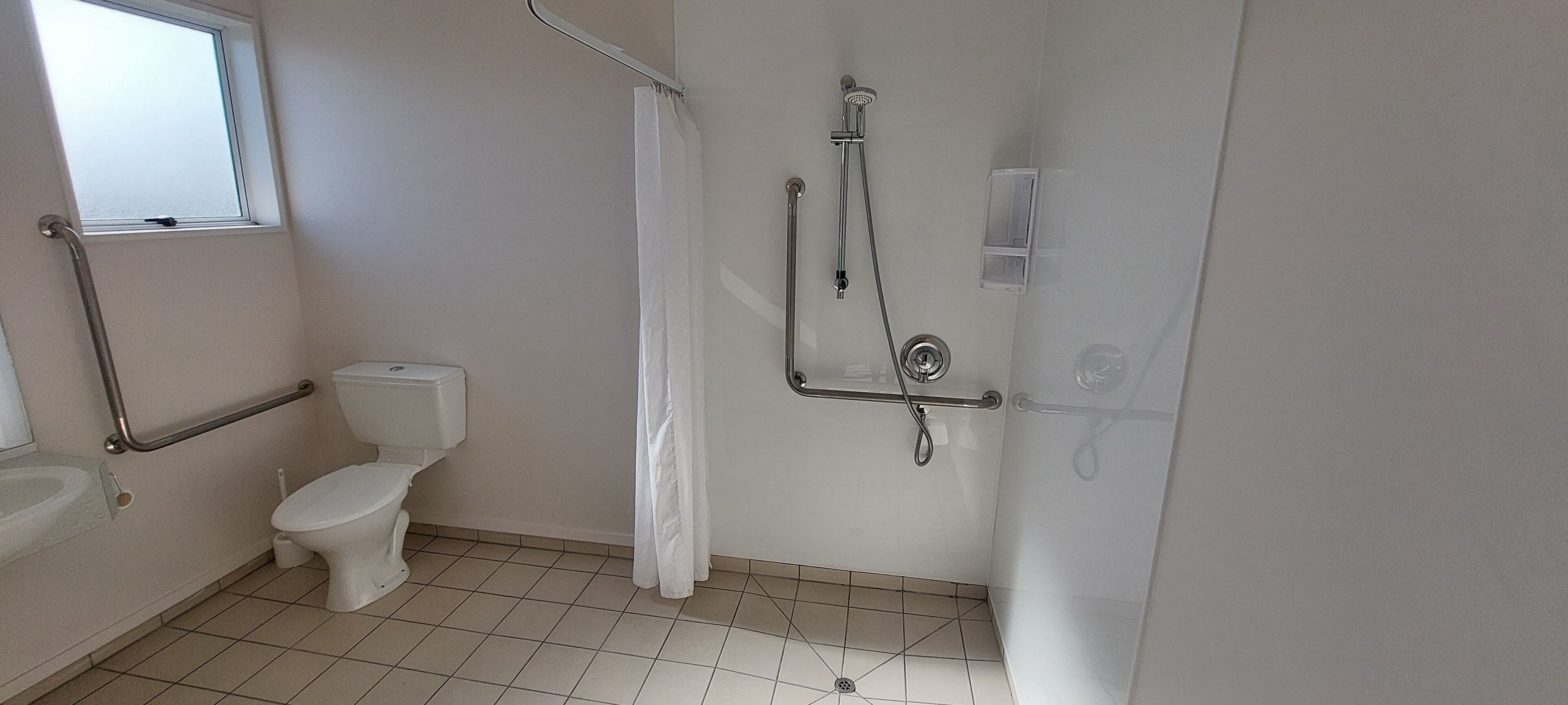 A bathroom with a toilet, a shower with a grab bar, a window, and a sink.
