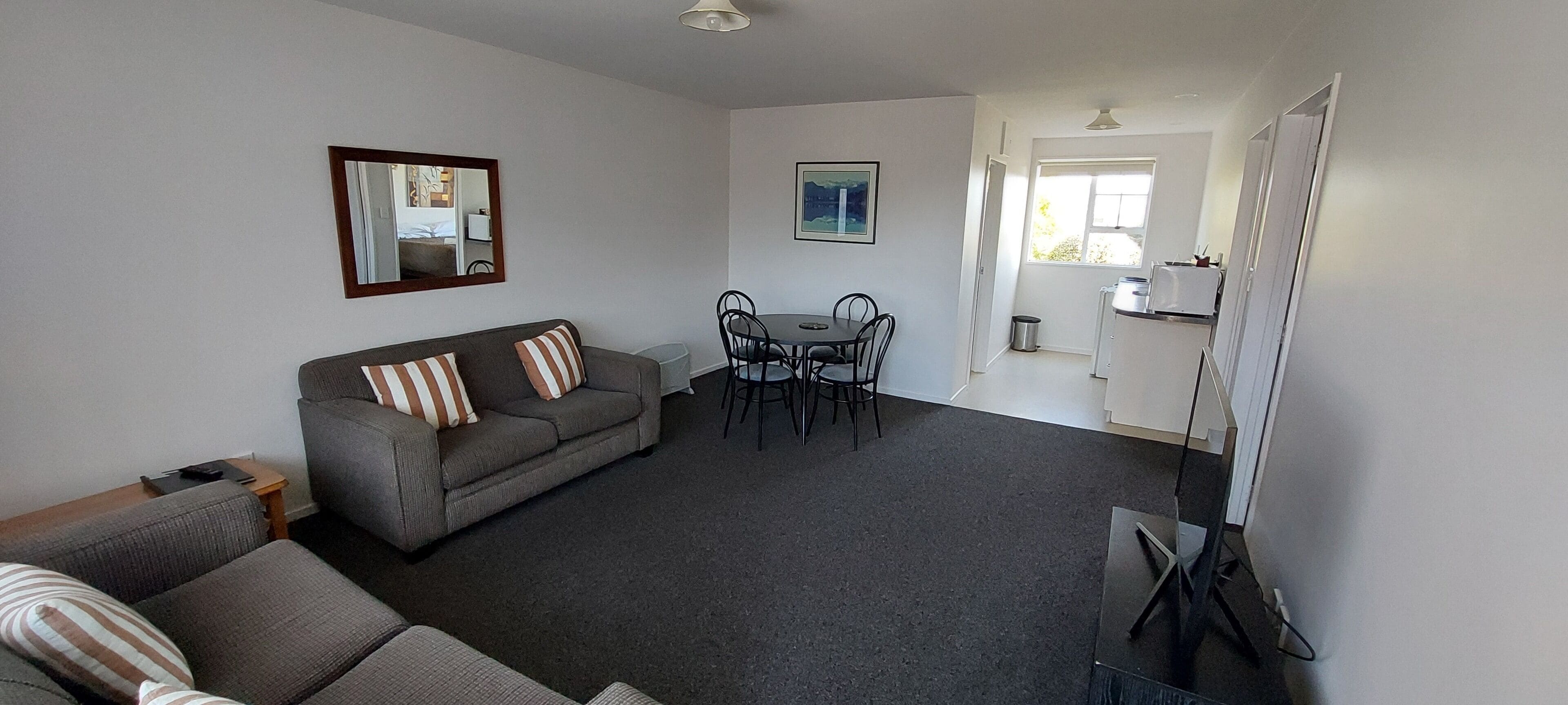 Ein Wohnzimmer mit grauem Sofa, gestreiften Kissen, einem Esstisch mit Stühlen und einer Küchenzeile mit Kühlschrank.