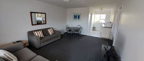 Ein Wohnzimmer mit grauem Sofa, gestreiften Kissen, einem Esstisch mit Stühlen und einer Küchenzeile mit Kühlschrank.