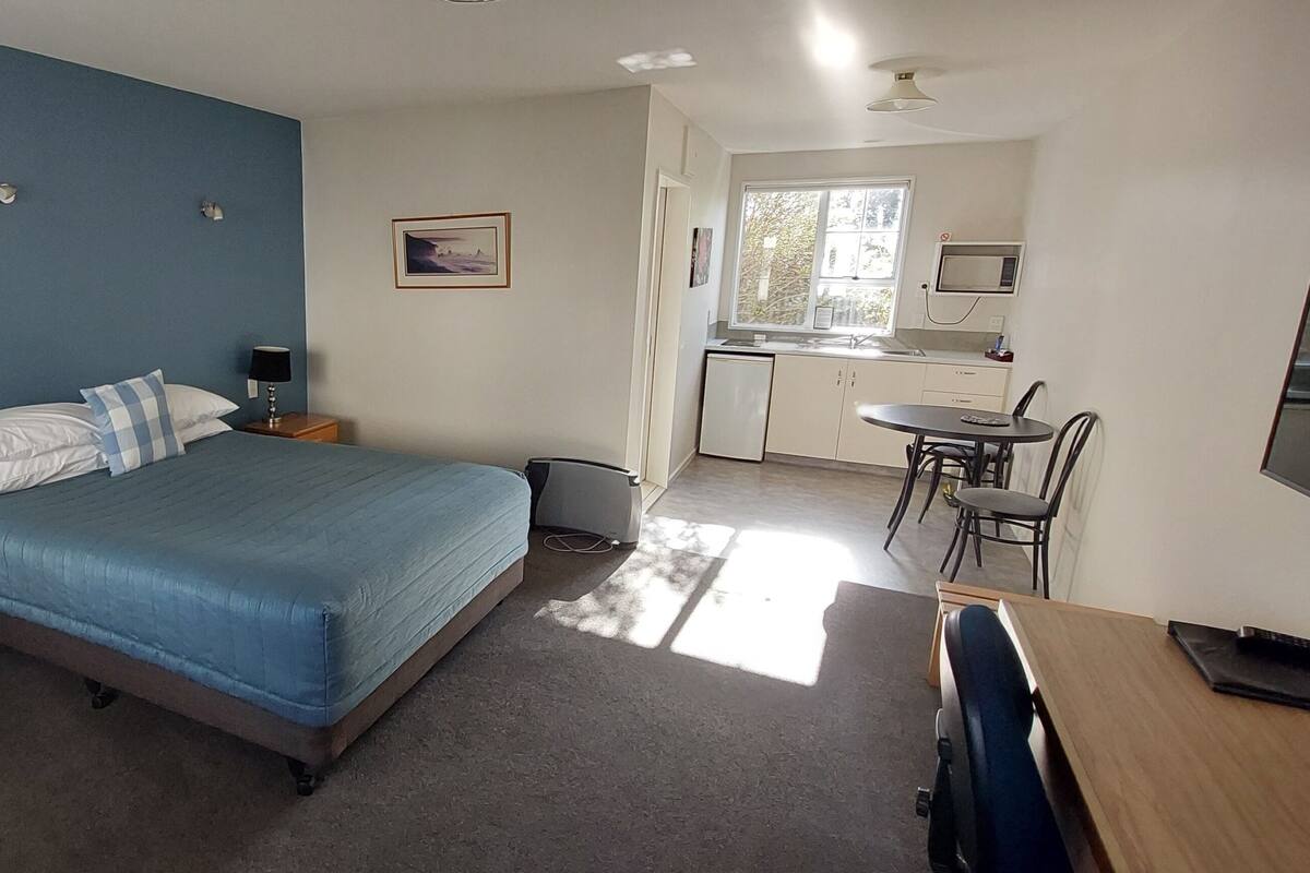 Quarto com cama, mesas de cabeceira, escrivaninha e cozinha compacta.