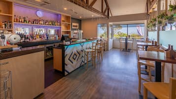 A bar area with a counter, stools, and a variety of bottles. There are tables and chairs in the dining area, and a large window with a view.