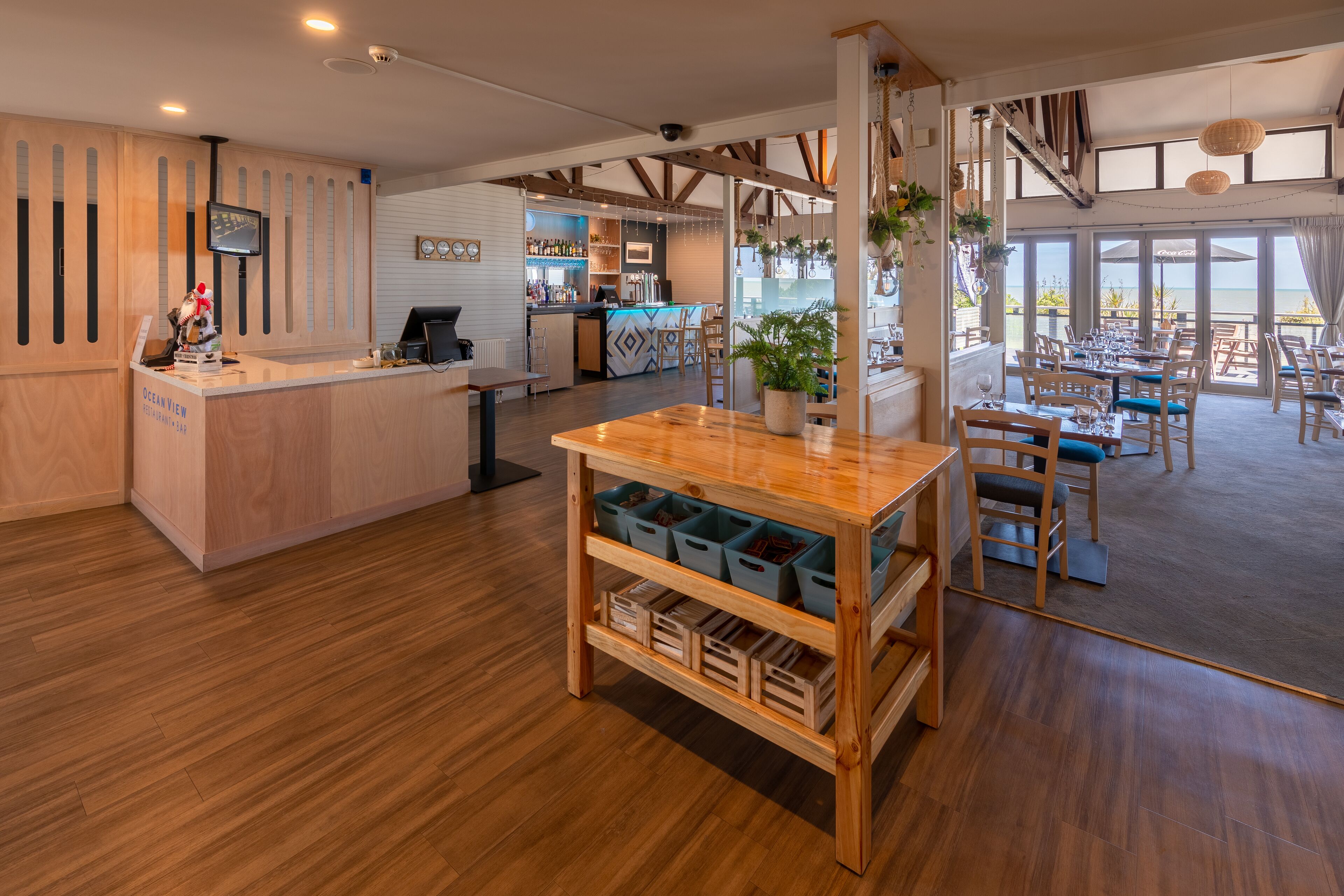 A spacious restaurant with wooden flooring, a bar area, and large windows offering a view of the outdoors.