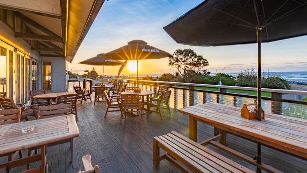 An outdoor patio with wooden tables and chairs, umbrellas, and a view of the sunset over the ocean. - Beachfront Hotel (Hokitika)