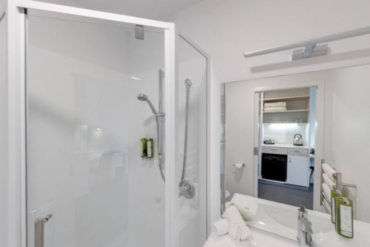 A modern bathroom with a glass shower enclosure, a white sink with a mirror, and a visible kitchen area in the background.