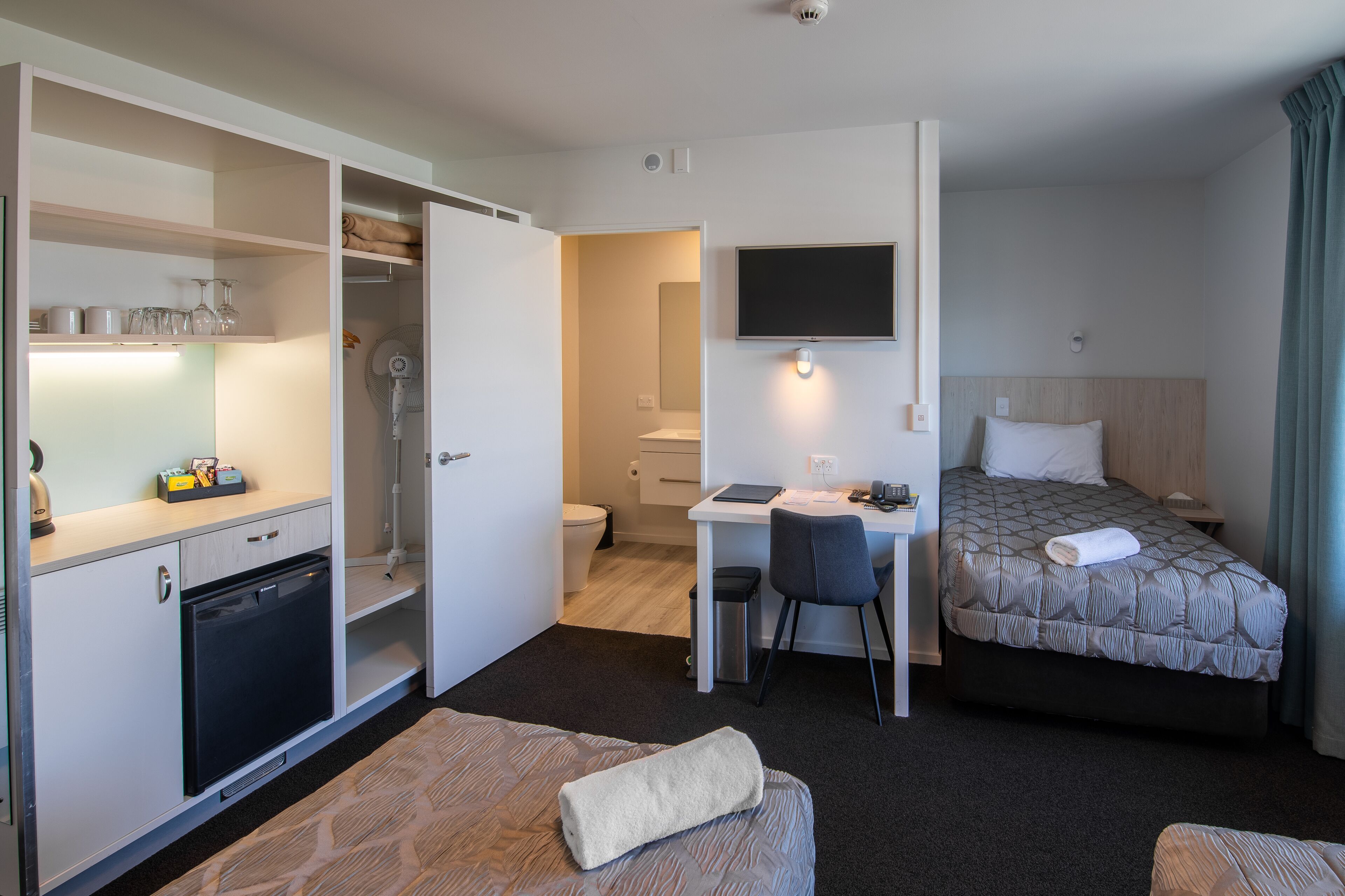 A hotel room with a bed, a desk with a chair, a small kitchenette, and a TV mounted on the wall.