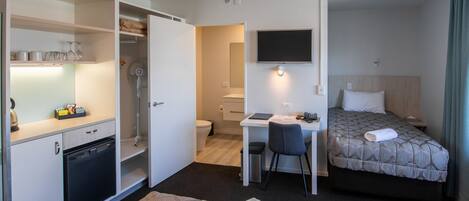 A hotel room with a bed, a desk with a chair, a small kitchenette, and a TV mounted on the wall.