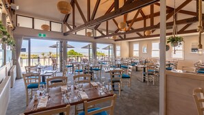 A spacious dining area with multiple tables and chairs, large windows with a view, and a high ceiling with exposed beams. - Beachfront Hotel (Hokitika)