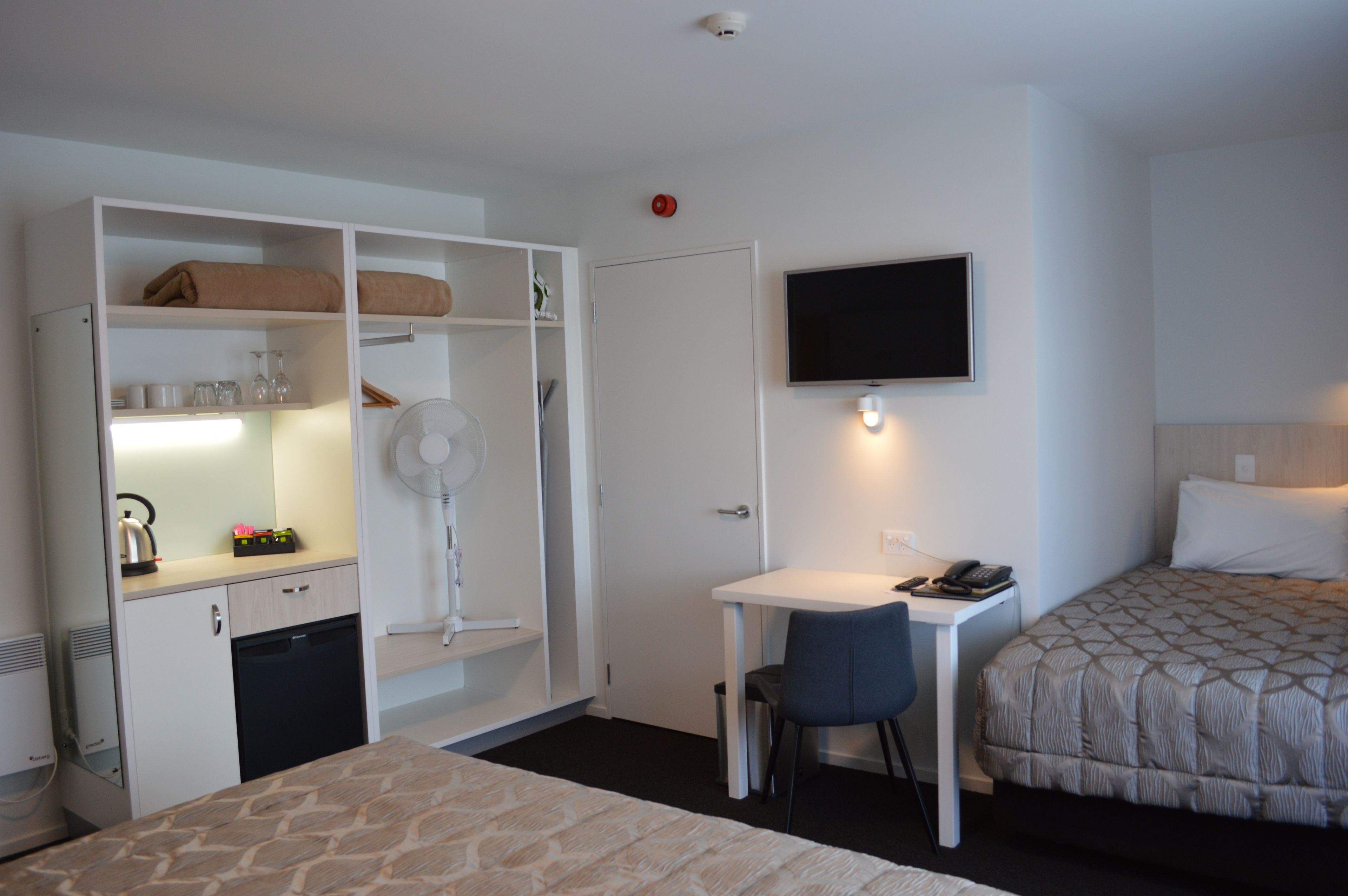 A hotel room with a bed, a desk with a chair, a TV, a wardrobe, and a small kitchenette.