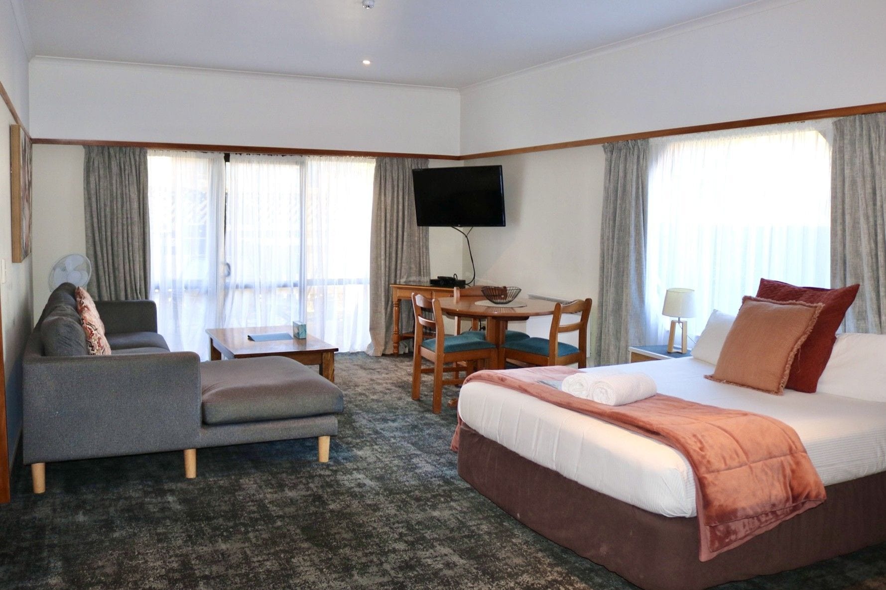 A hotel room with a bed, a sofa, a desk with chairs, a television, and large windows with curtains.