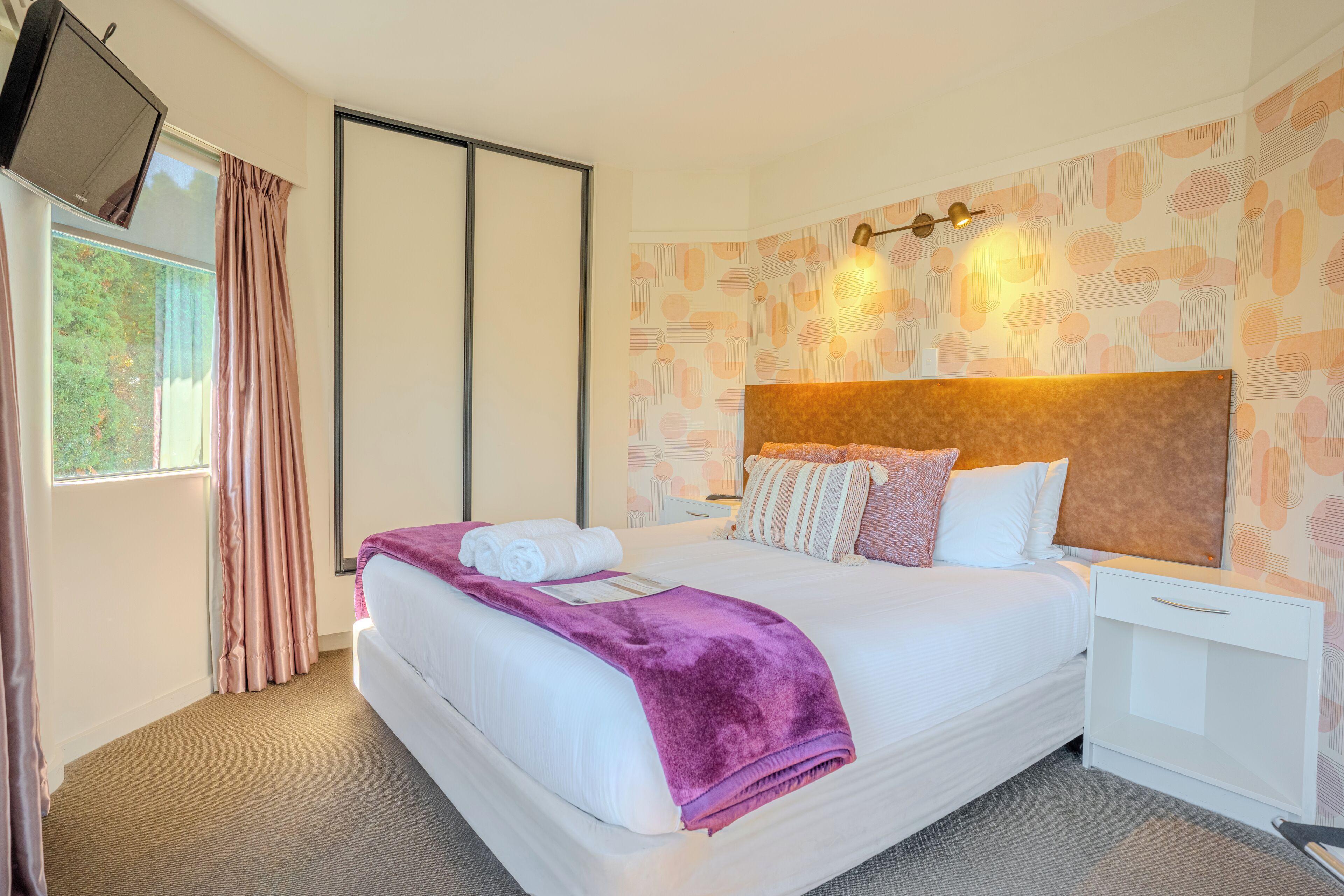 Une chambre d’hôtel avec un lit, une table de chevet, un téléviseur fixé au mur et une fenêtre avec des rideaux.