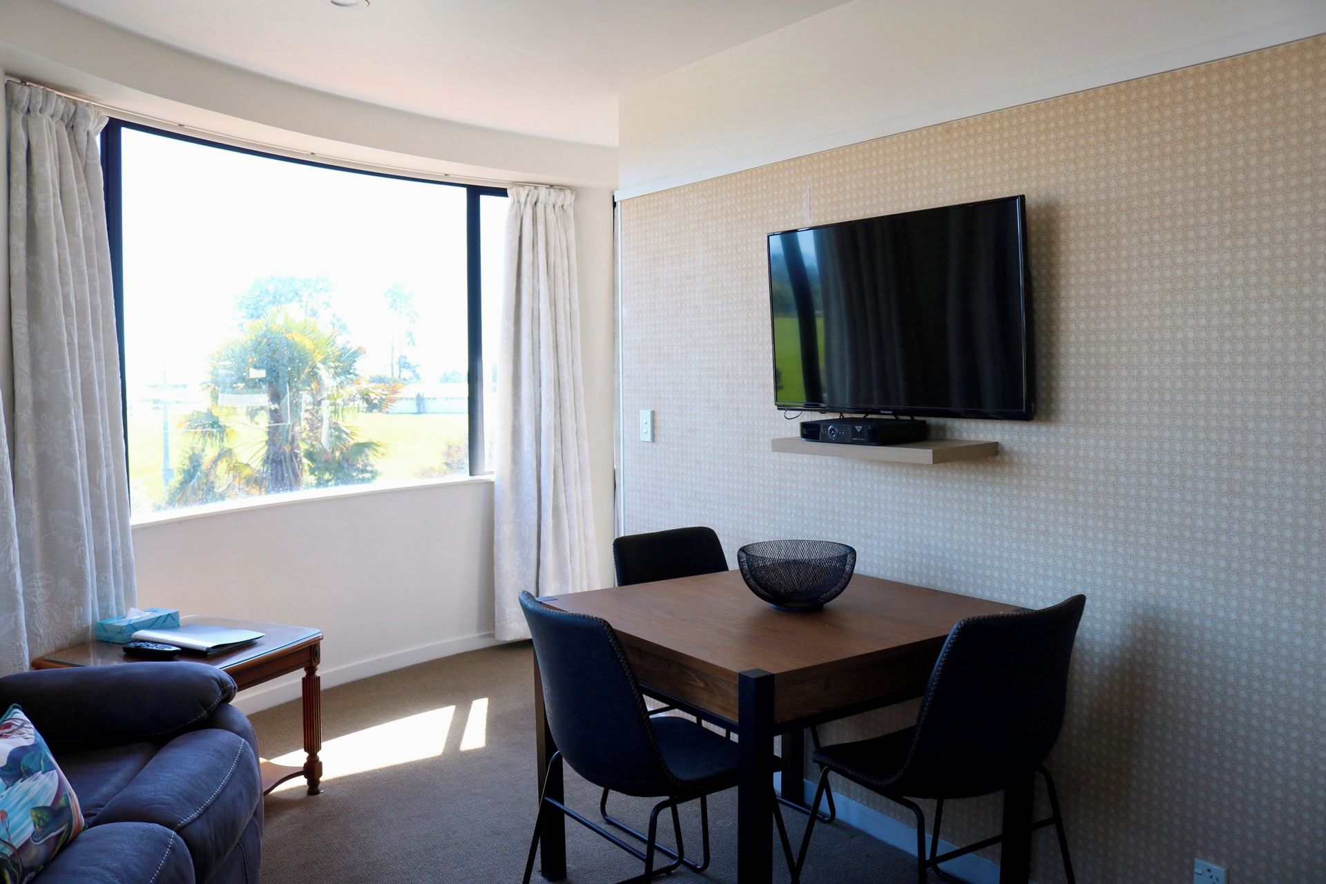 A hotel room with a dining table, chairs, a TV mounted on the wall, and a large window with curtains.