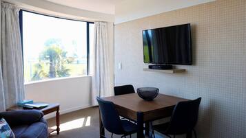 Habitación de hotel con una mesa de comedor, sillas, un televisor montado en la pared y un amplio ventanal con cortinas.