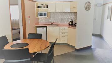 Una cocina compacta con microondas, hervidor y un refrigerador pequeño. Hay una mesa de comedor con sillas y un reloj en la pared.