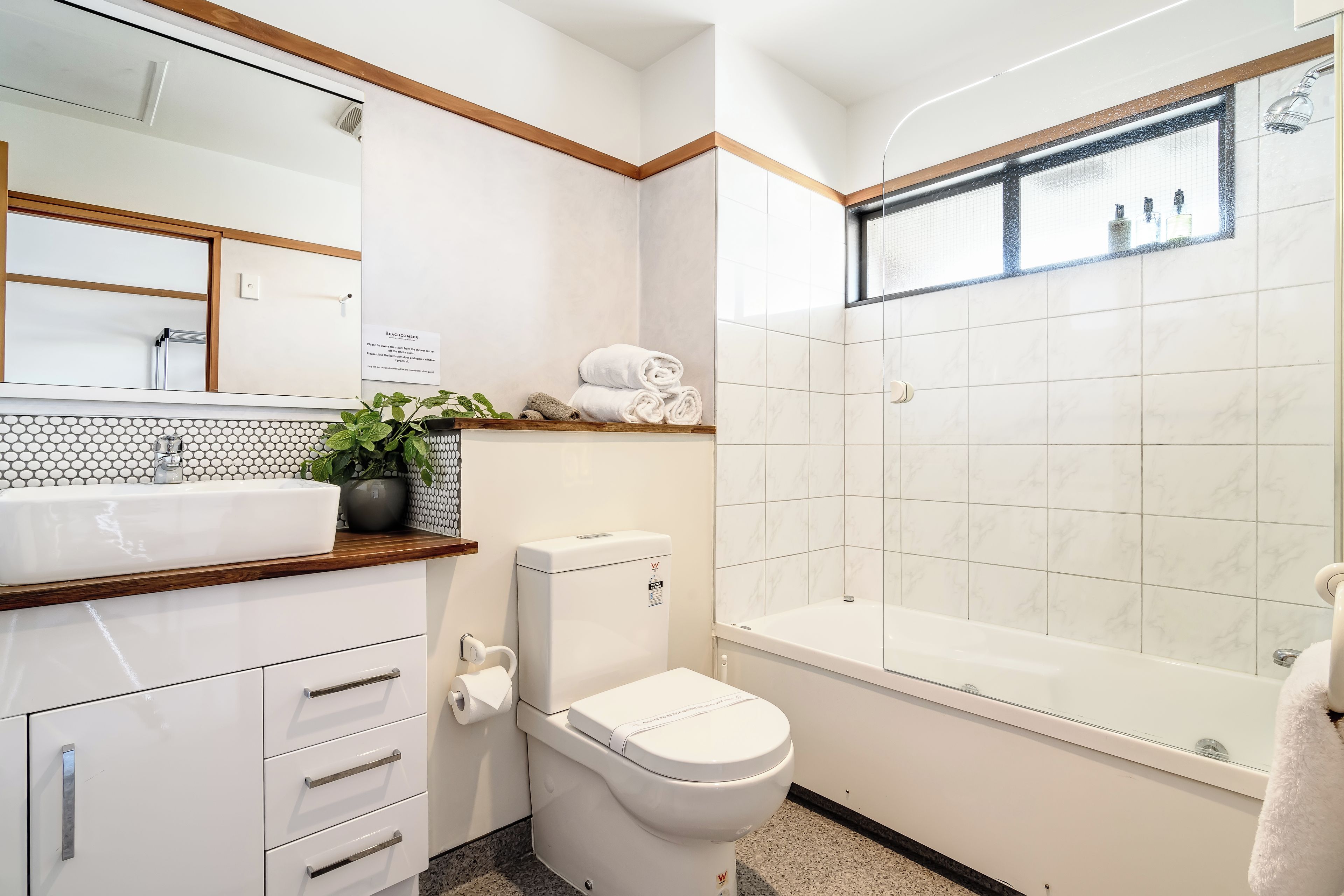 A modern bathroom with a white sink, toilet, and bathtub. There is a mirror above the sink and a shelf with towels.