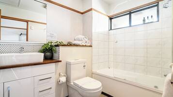 A modern bathroom with a white sink, toilet, and bathtub. There is a mirror above the sink and a shelf with towels.