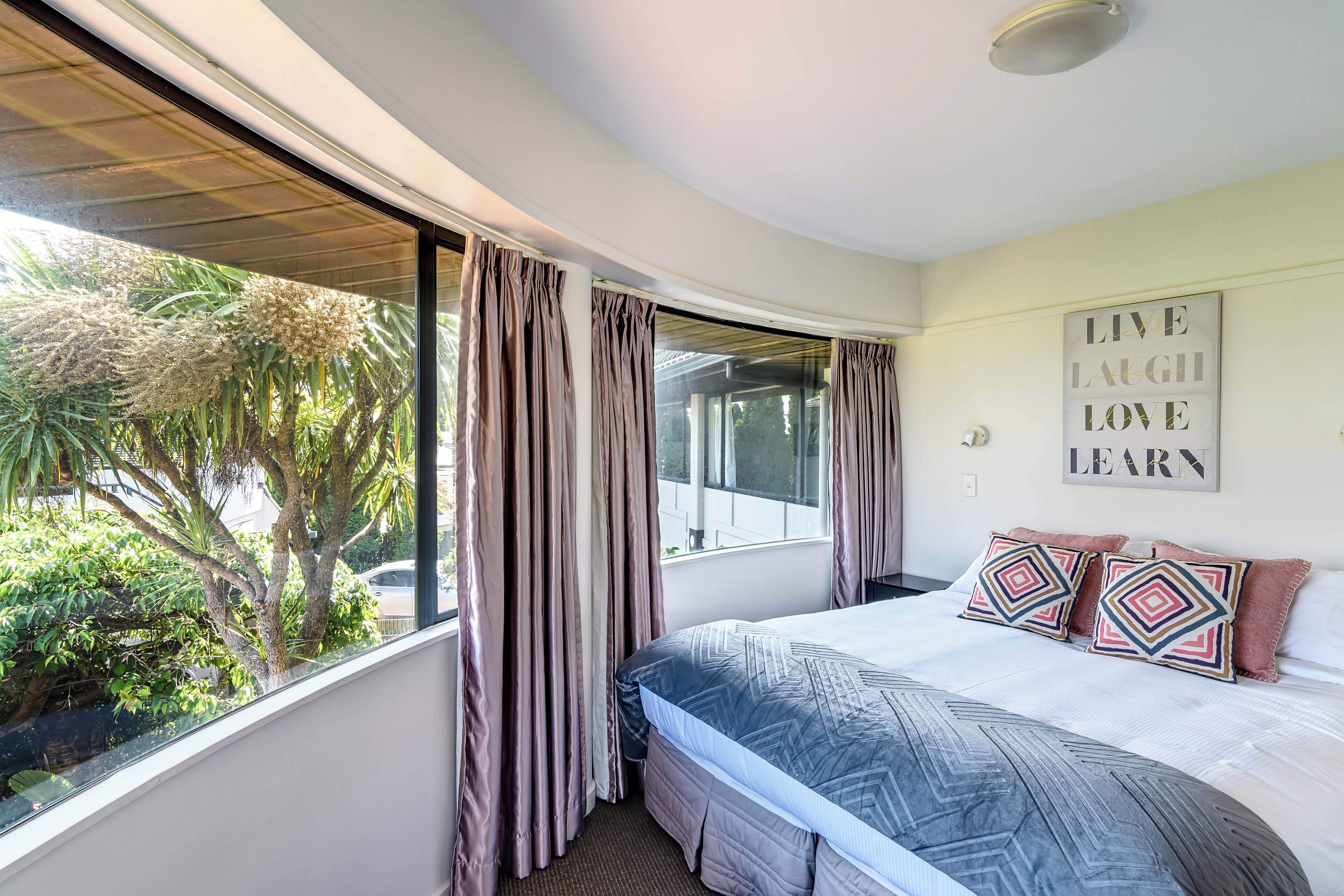 A bedroom with a large window, a bed with patterned bedding, and a motivational wall quote.