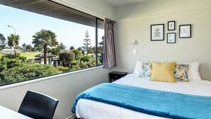 A hotel room with a bed, a desk, a chair, a window with a view of a golf course, and framed pictures on the wall. - The Beachcomber Hotel (Nelson)