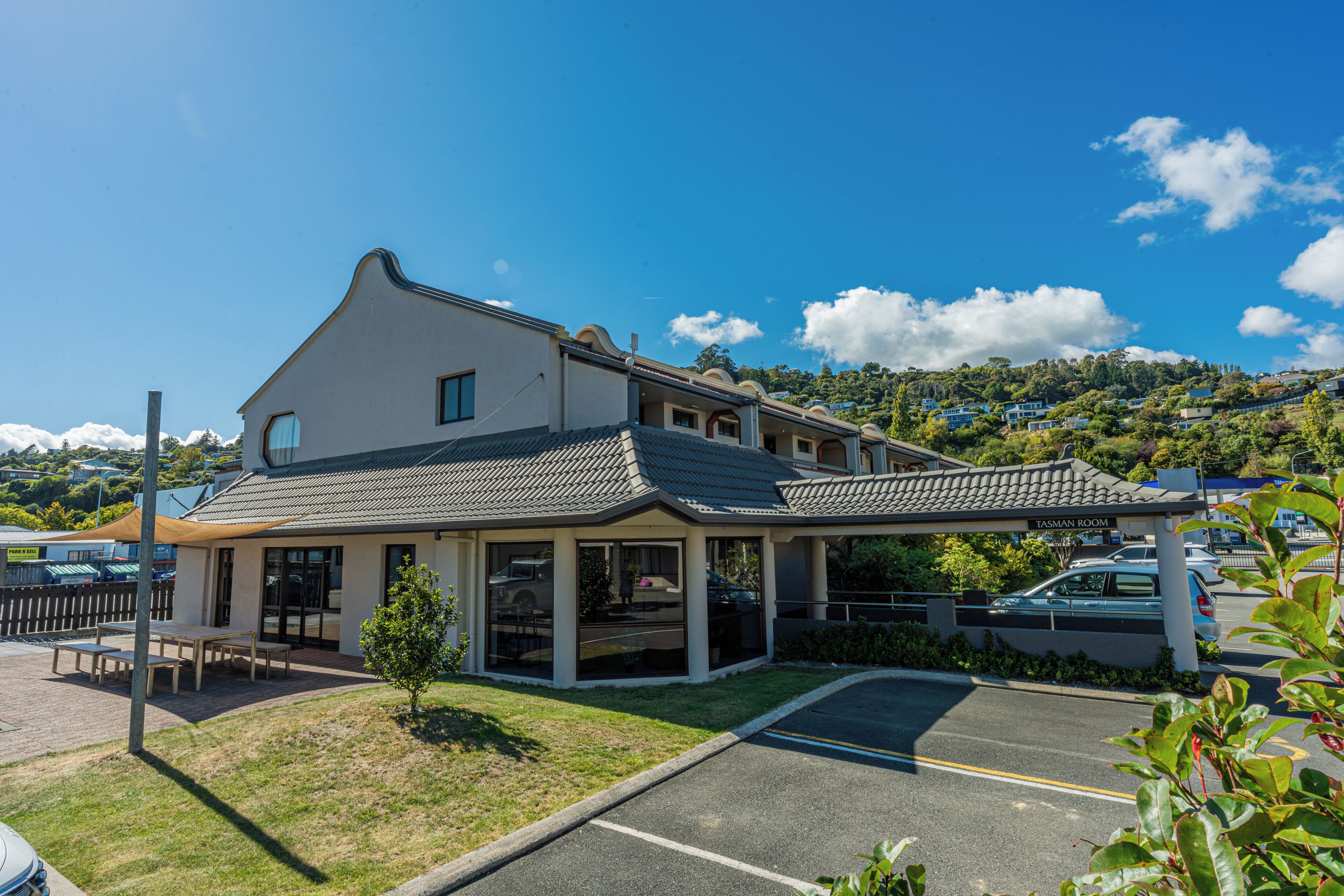 A hotel with a covered outdoor seating area, a parking lot, and a view of a hilly landscape.