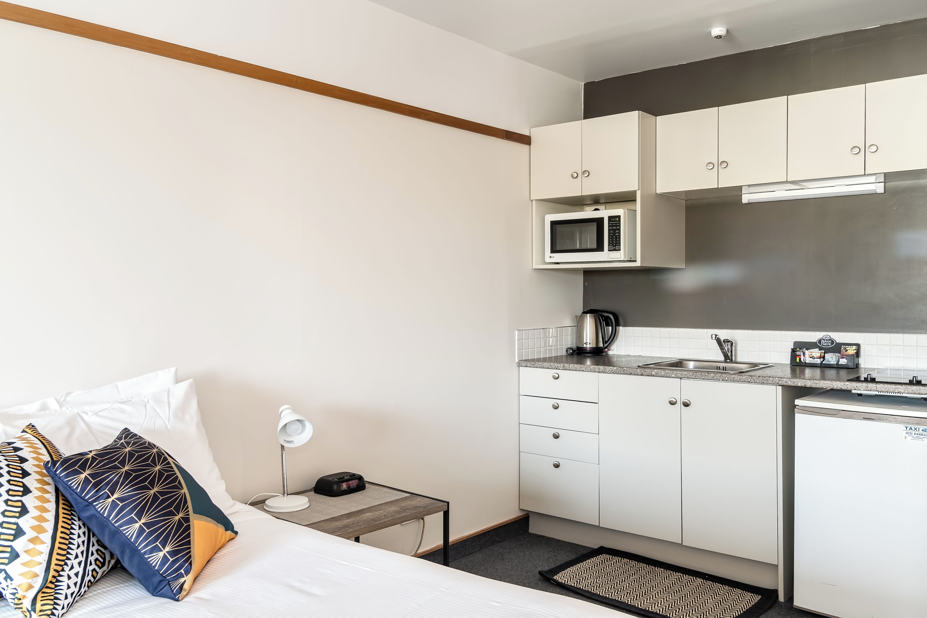 Habitación de hotel compacta con cama, mesita de noche, lámpara y una kitchenette equipada con microondas, tetera y refrigerador pequeño.