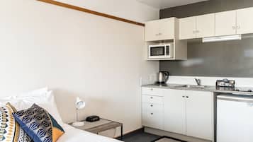 Habitación de hotel compacta con cama, mesita de noche, lámpara y una kitchenette equipada con microondas, tetera y refrigerador pequeño.
