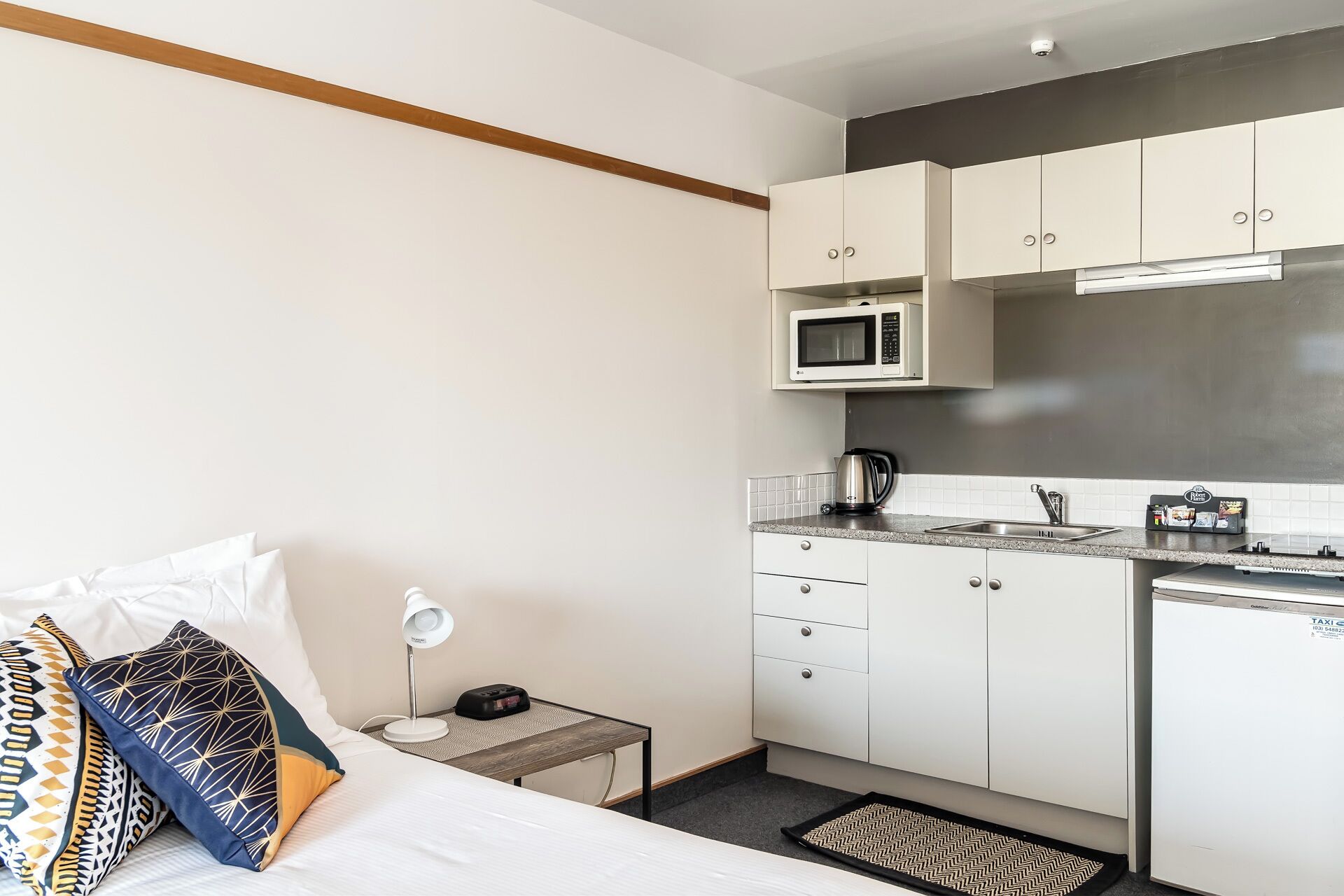 Een kleine hotelkamer met een bed, nachtkastje, lamp en een kitchenette met een magnetron, waterkoker en spoelbak.
