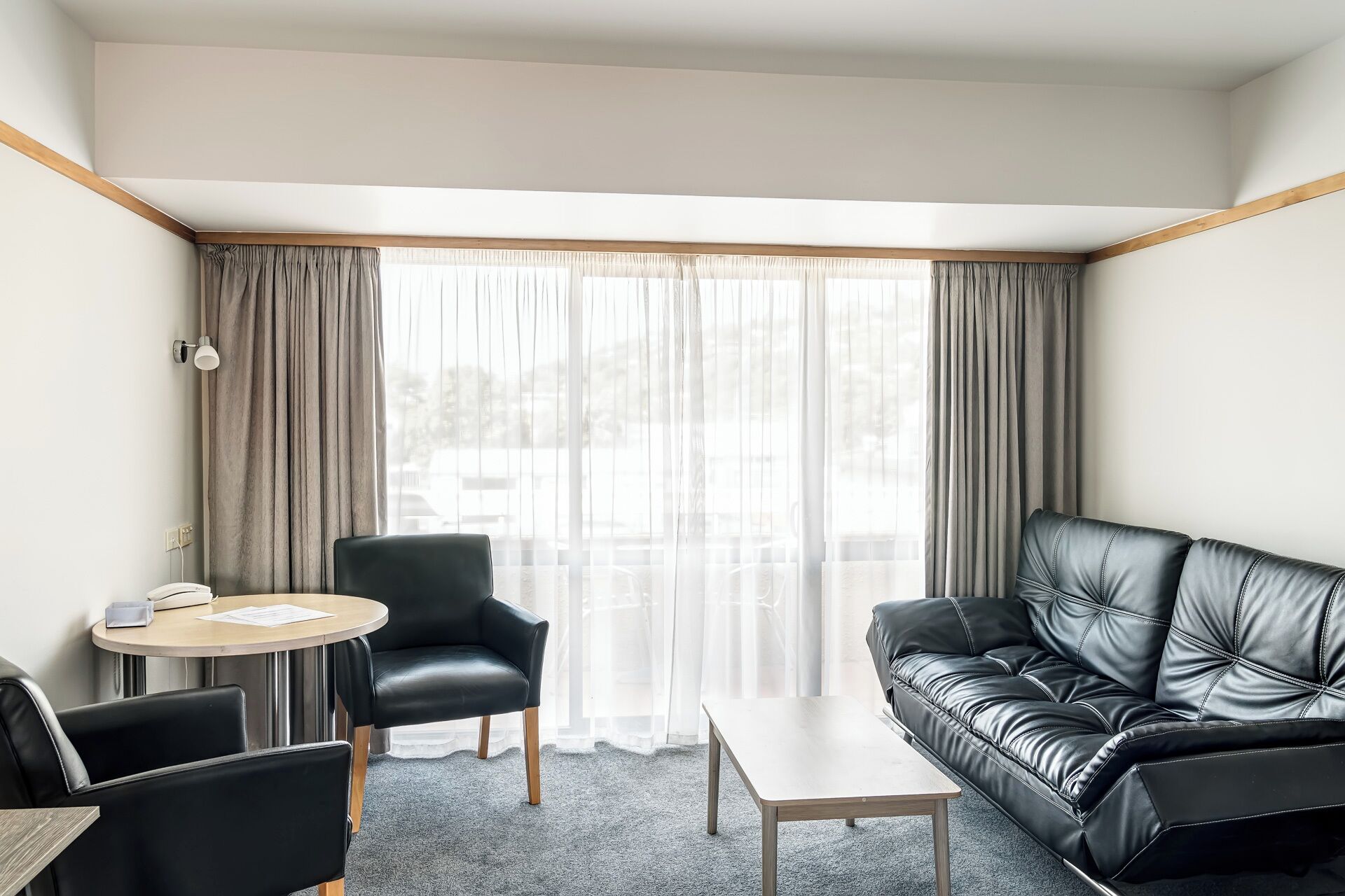A hotel room with a black leather sofa, a round table, a chair, and a large window with curtains.