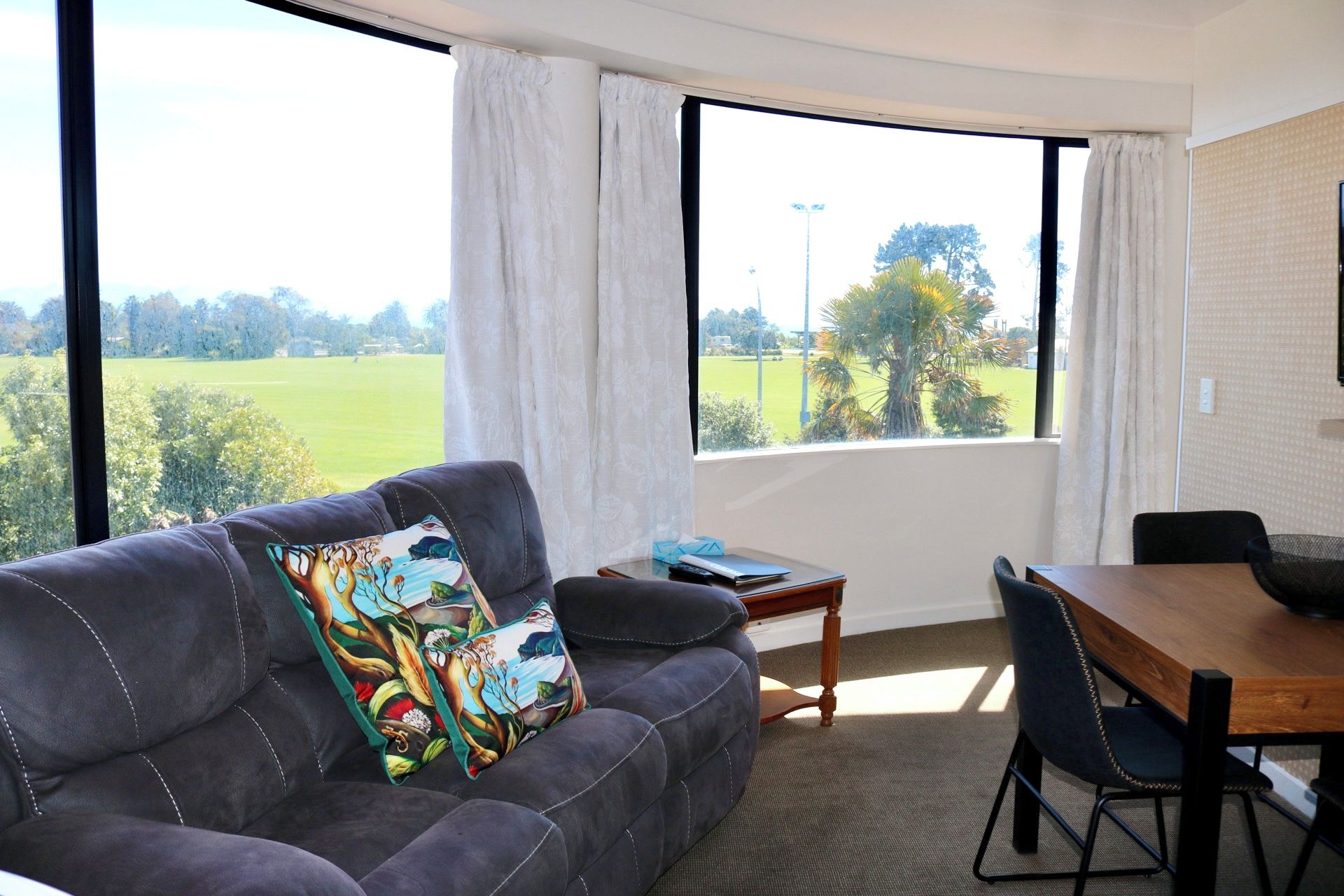 A room with a grey sofa, a wooden table, and a view of a green field through large windows.