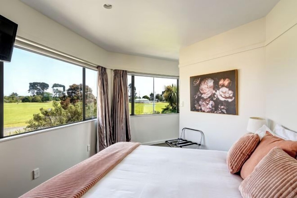 A bedroom with a large window, a bed with pink bedding, a painting on the wall, and a chair.
