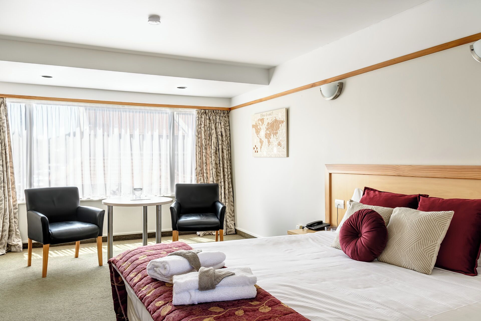 A hotel room with a bed, two chairs, a small table, a window with curtains, and a wall-mounted light.