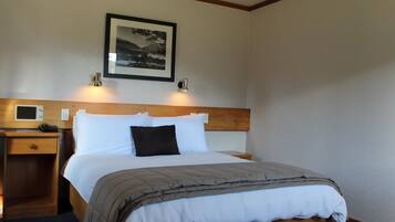 Habitación de hotel con cabecera de madera, cama con ropa de cama blanca y gris, mesita de noche con lámpara y un cuadro en la pared.