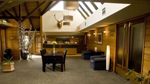 Un vestíbulo de hotel con mostrador de recepción, zona de comedor con mesa y sillas, sala de estar con sofás y una gran pared de madera con un ciervo disecado.