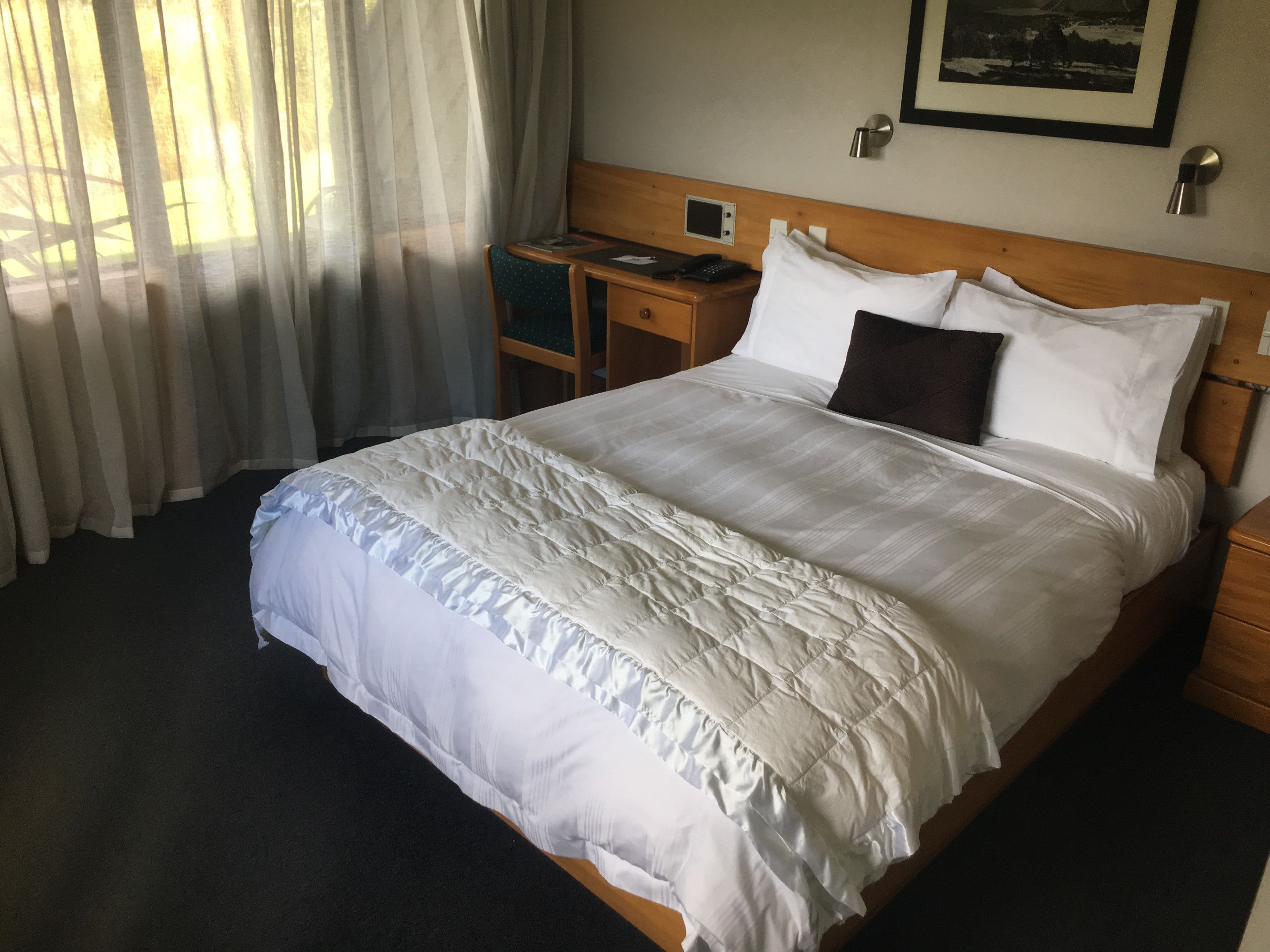 A neatly made bed with a white duvet and pillows, a bedside table with a phone and a small electronic device, and a window with sheer curtains allowing natural light.