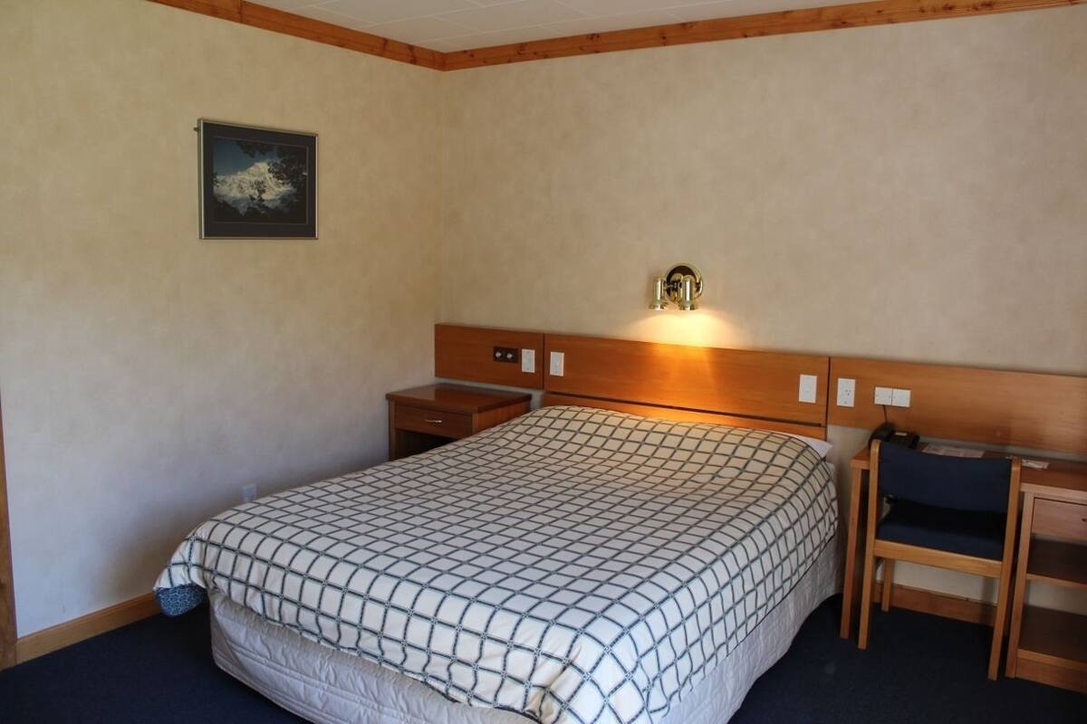 A hotel room with a large bed, a nightstand, a desk with a chair, and a wall-mounted light fixture.