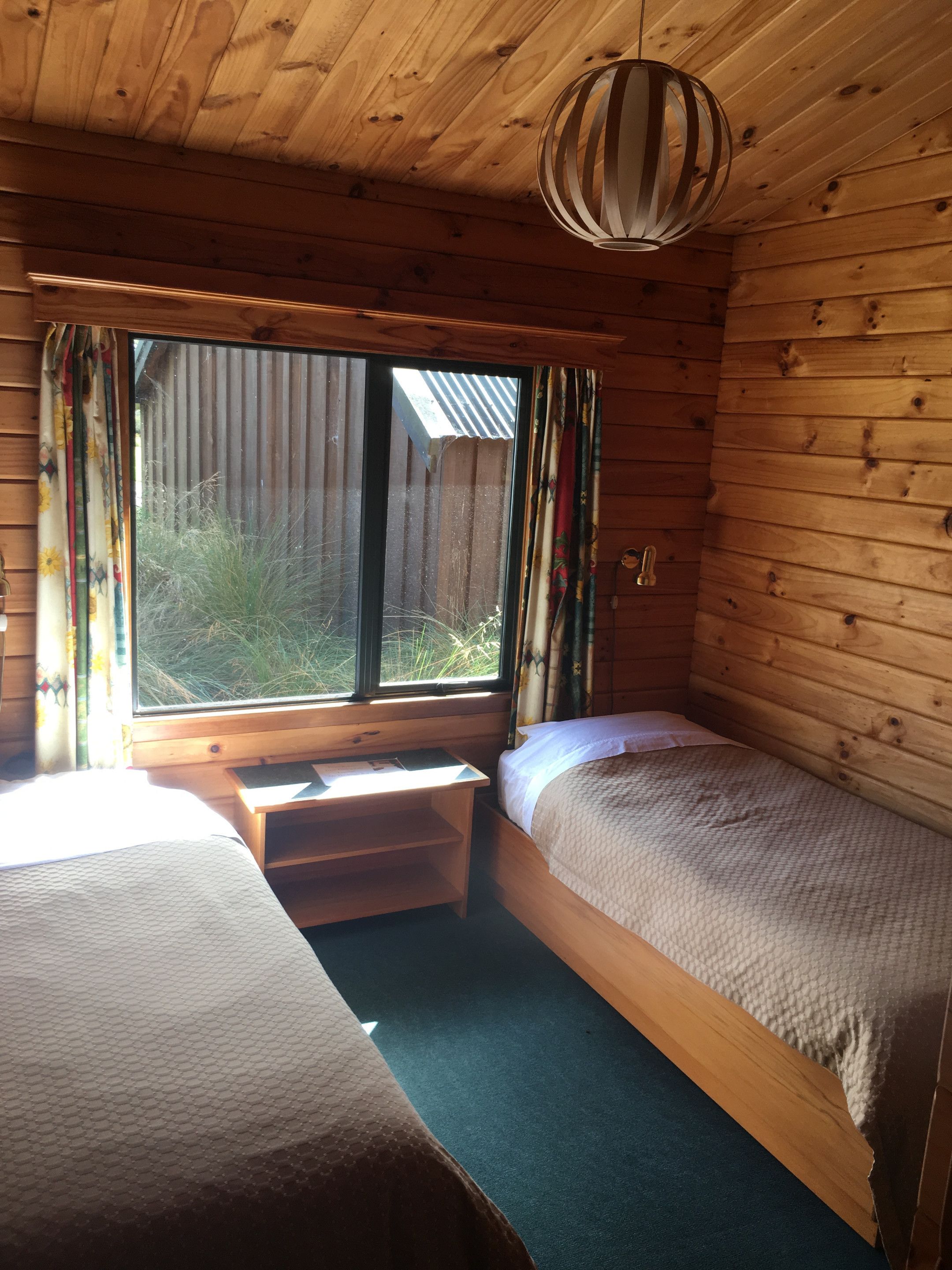 A cabin room with two beds, a wooden ceiling, a window with curtains, and a hanging light fixture.