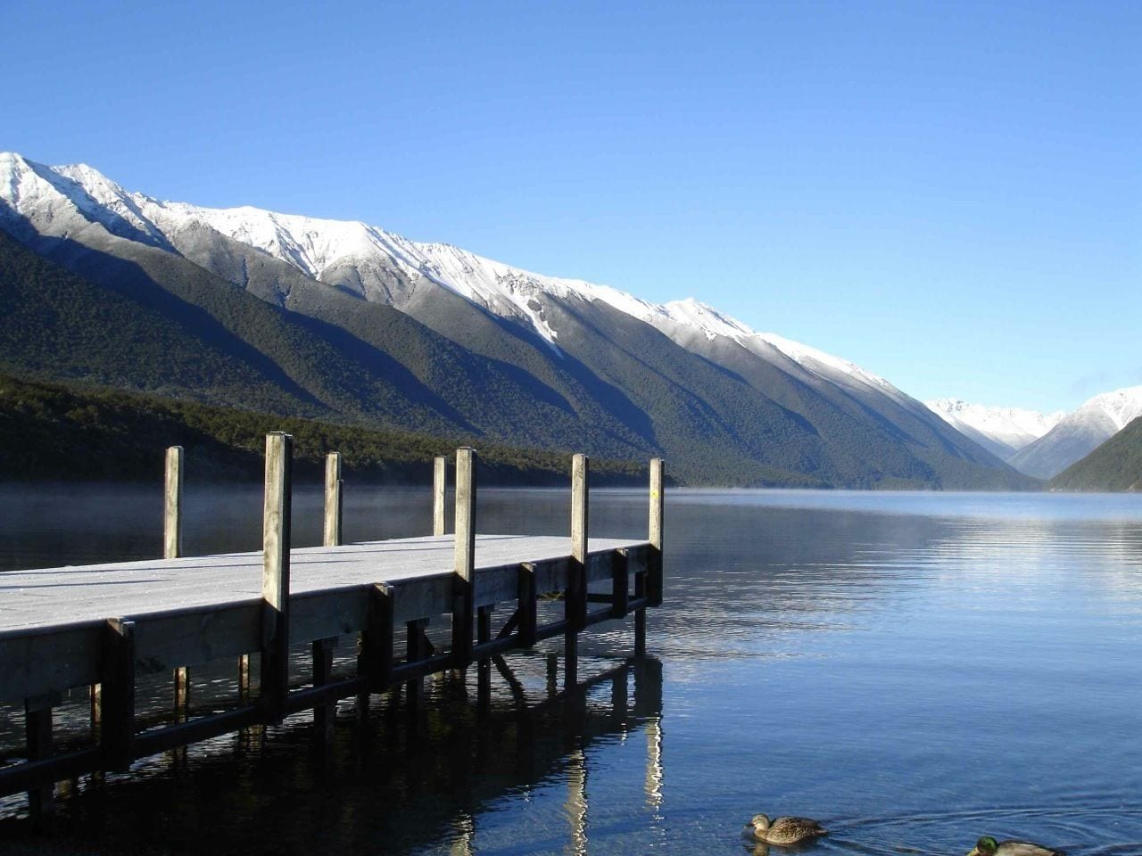 A wooden dock extending into a calm lake with snow-capped mountains in the background.