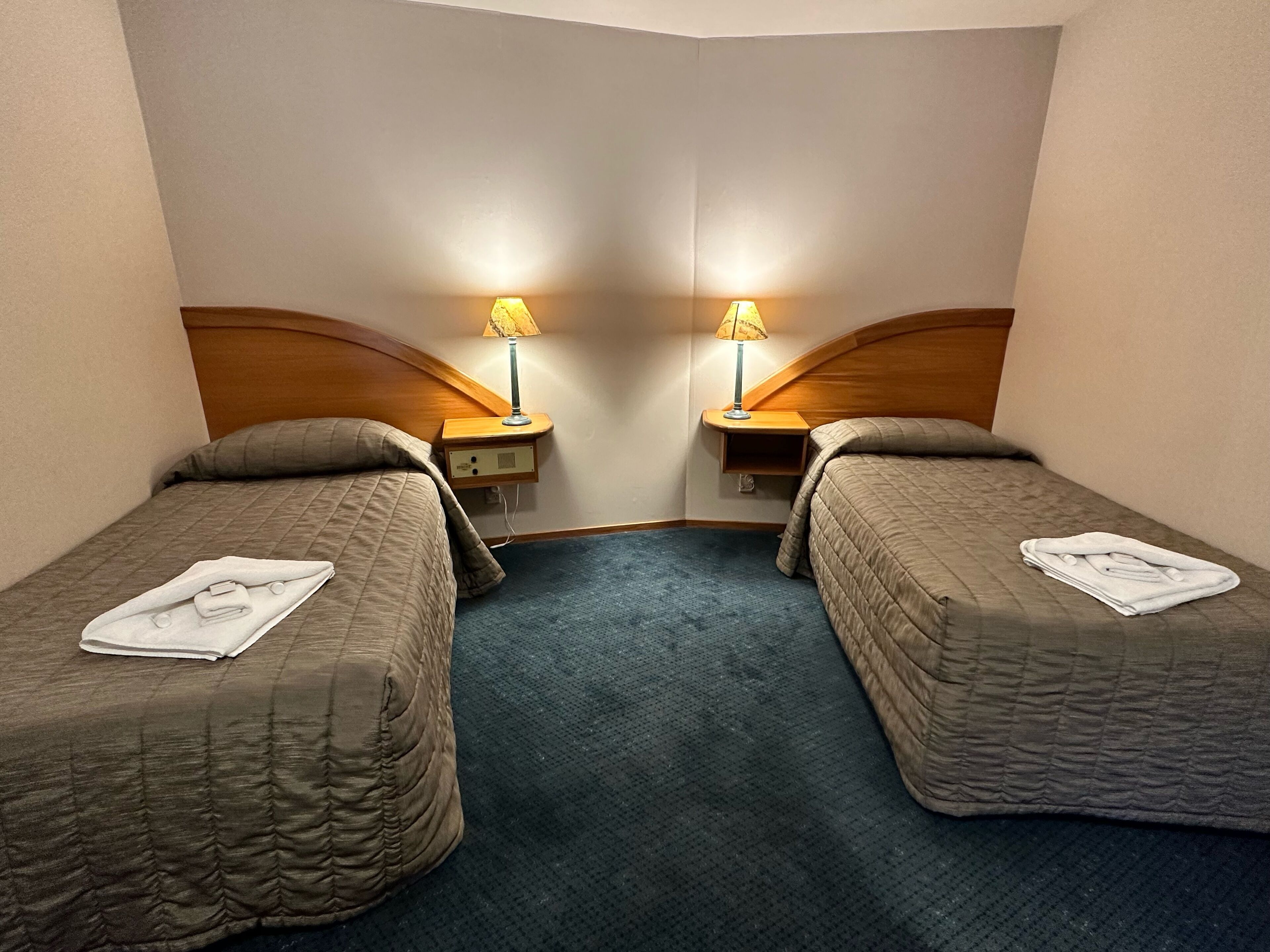 A hotel room with two single beds, each with a pillow and a folded towel. There are bedside tables with lamps and a small shelf. The room has a carpeted floor and a wooden headboard.