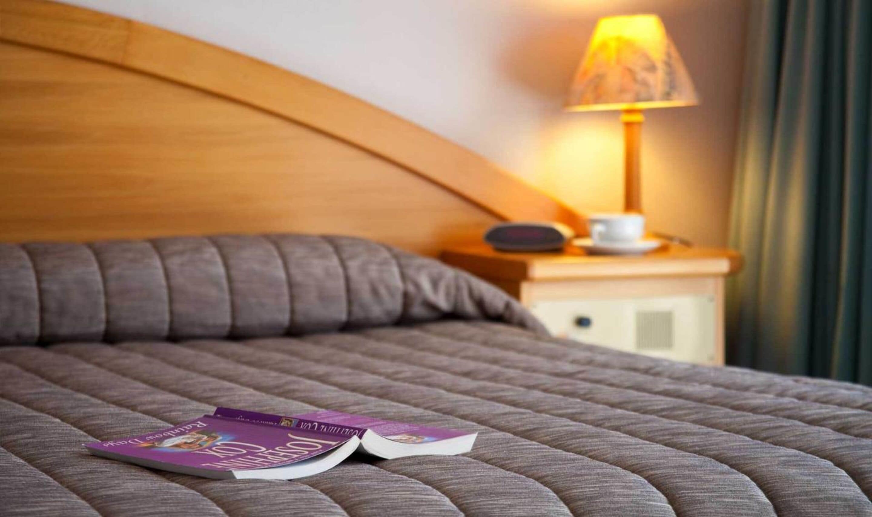 Una cama bien hecha con cabecera, una mesita de noche con lámpara y una revista sobre la cama.