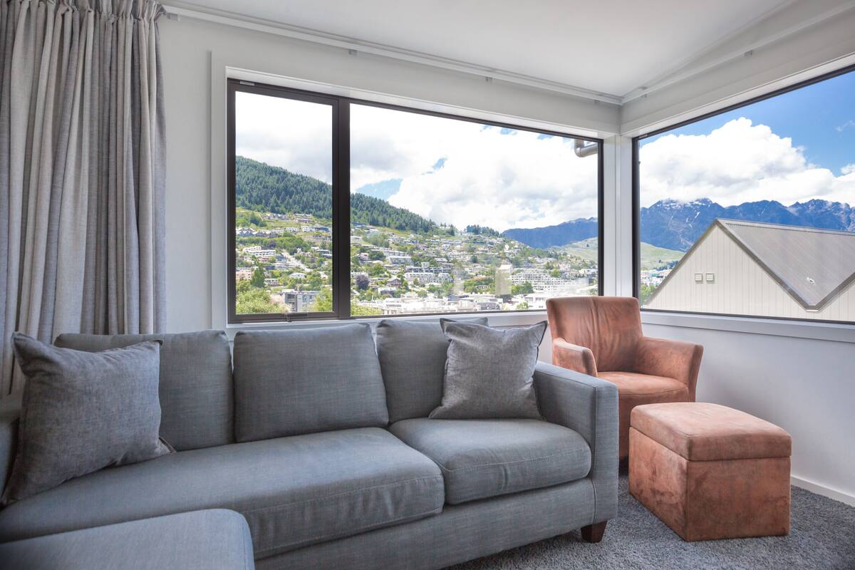 Ein modernes Wohnzimmer mit einem grauen Sofa, einem braunen Sessel und einem großen Fenster mit Blick auf eine Stadt und Berge.