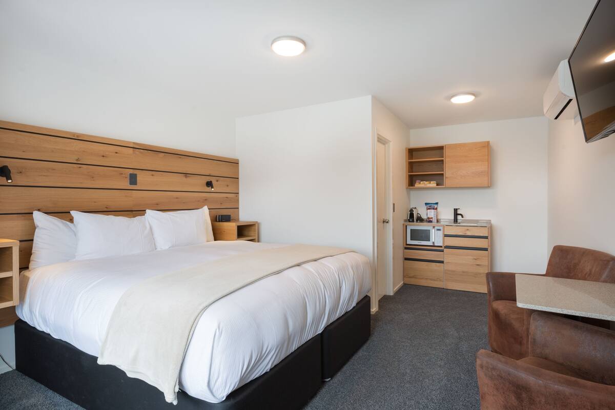 Ein modernes Hotelzimmer mit einem großen Bett, einem Holz-Kopfteil, einem Flachbildfernseher, einer Mikrowelle und einer kleinen Küchenzeile.