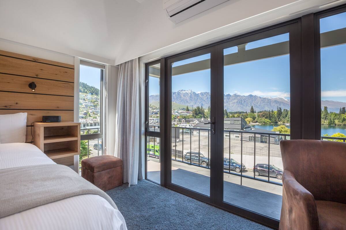 Ein Hotelzimmer mit großem Fenster, einem Bett, einem hölzernen Kopfteil, einem kleinen Tisch und einem Stuhl.