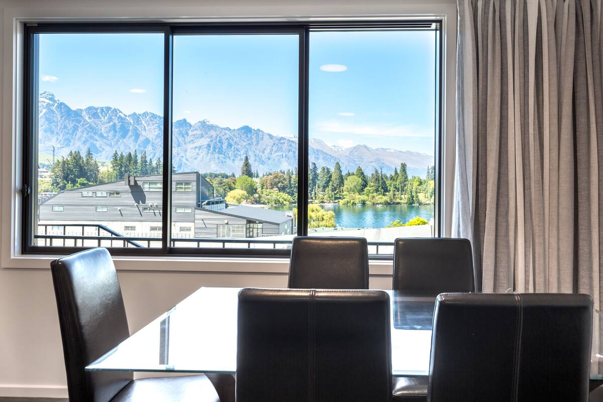 Ein Zimmer mit großem Fenster und Blick auf Berge und einen See. Vor dem Fenster steht ein Tisch mit Stühlen.