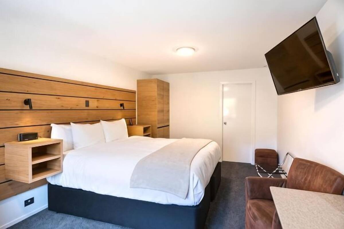 Ein modernes Hotelzimmer mit einem großen Bett, einem an der Wand befestigten Fernseher, einem Holz-Kopfteil und einer kleinen Sitzecke.