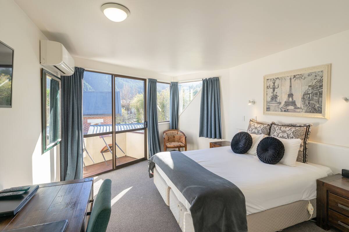 A hotel room with a large bed, a desk, and a chair. There is a balcony with a view of the city.