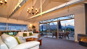 Un vaste espace de vie agrémenté d’un canapé blanc, d’une table basse en bois et d’une cheminée. Une grande fenêtre offre une vue sur les montagnes et donne accès à une terrasse dotée de mobilier d’extérieur.