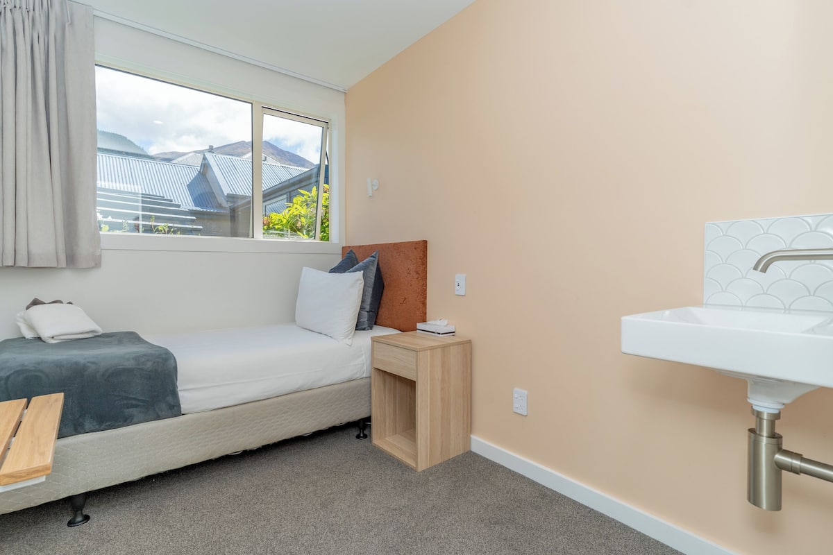 A bedroom with a single bed, a bedside table, a window with curtains, and a bathroom with a sink.