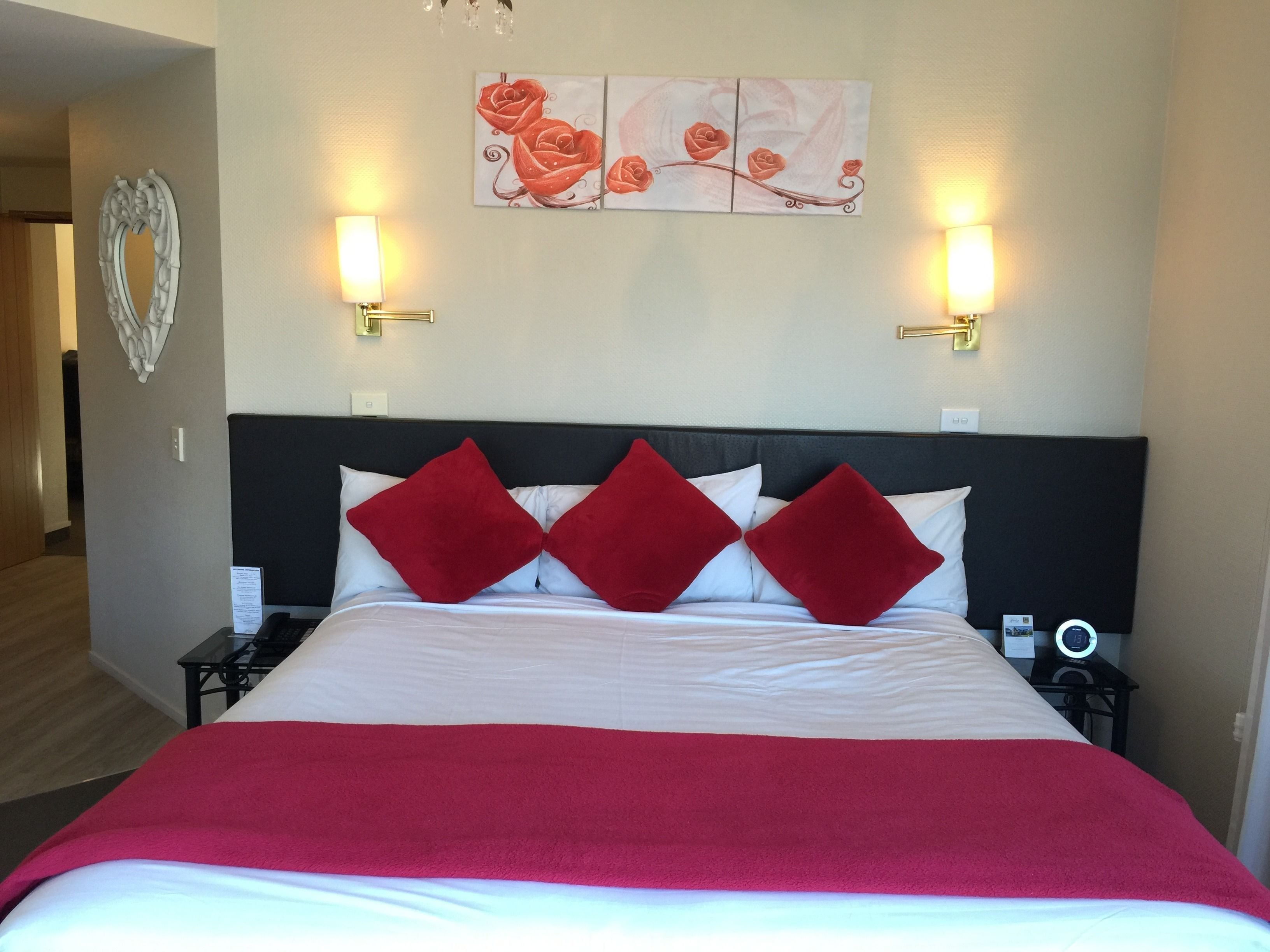 Una habitación de hotel con una cama grande, almohadas rojas y blancas, una manta roja y dos mesitas de noche con lámparas, además de una obra de arte en la pared sobre la cama.
