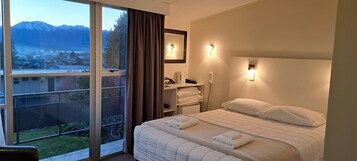 Habitación de hotel con un gran ventanal, una cama con ropa de cama blanca, una mesita de noche con lámpara y vistas a las montañas.