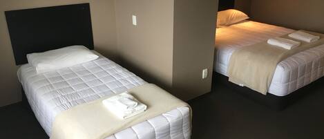 Two single beds with white bedding and a beige throw blanket, a dark headboard, and a wall-mounted light fixture.