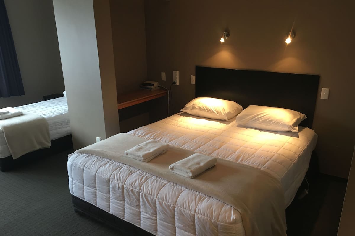 Una habitación de hotel con una cama grande, dos camas más pequeñas, una mesita de noche y una lámpara fijada a la pared.