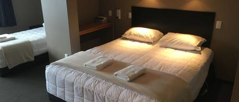 A hotel room with a large bed, two smaller beds, a bedside table, and a wall-mounted light fixture.